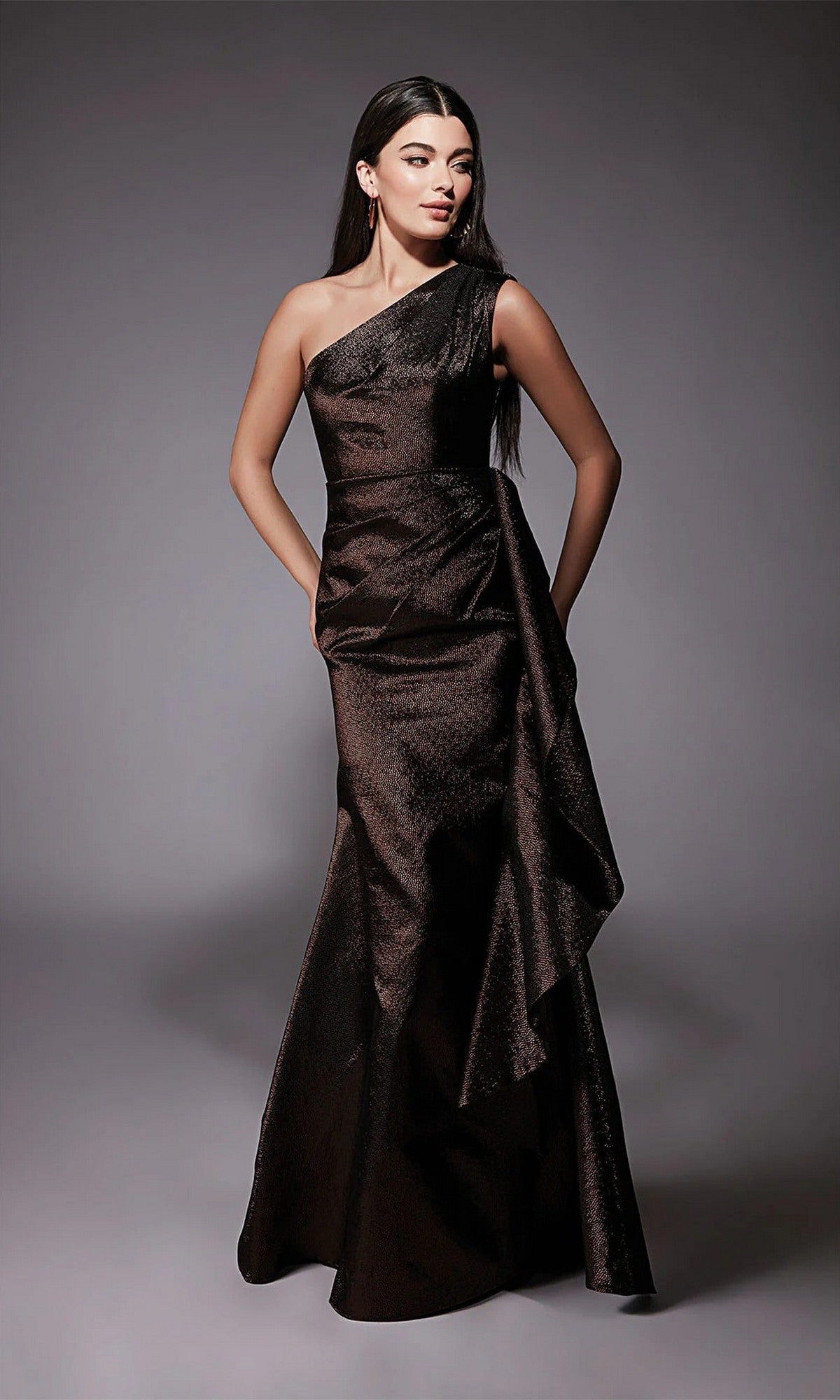 A woman wearing the Alyce 27751 long formal dress in brown, featuring a dark one-shoulder jacquard design, stands with hands on hips against a plain gray background.